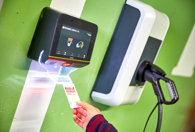 Ticket wird an einen Automaten gehalten. Im Hintergrund eine Ladesäule für E-Autos.